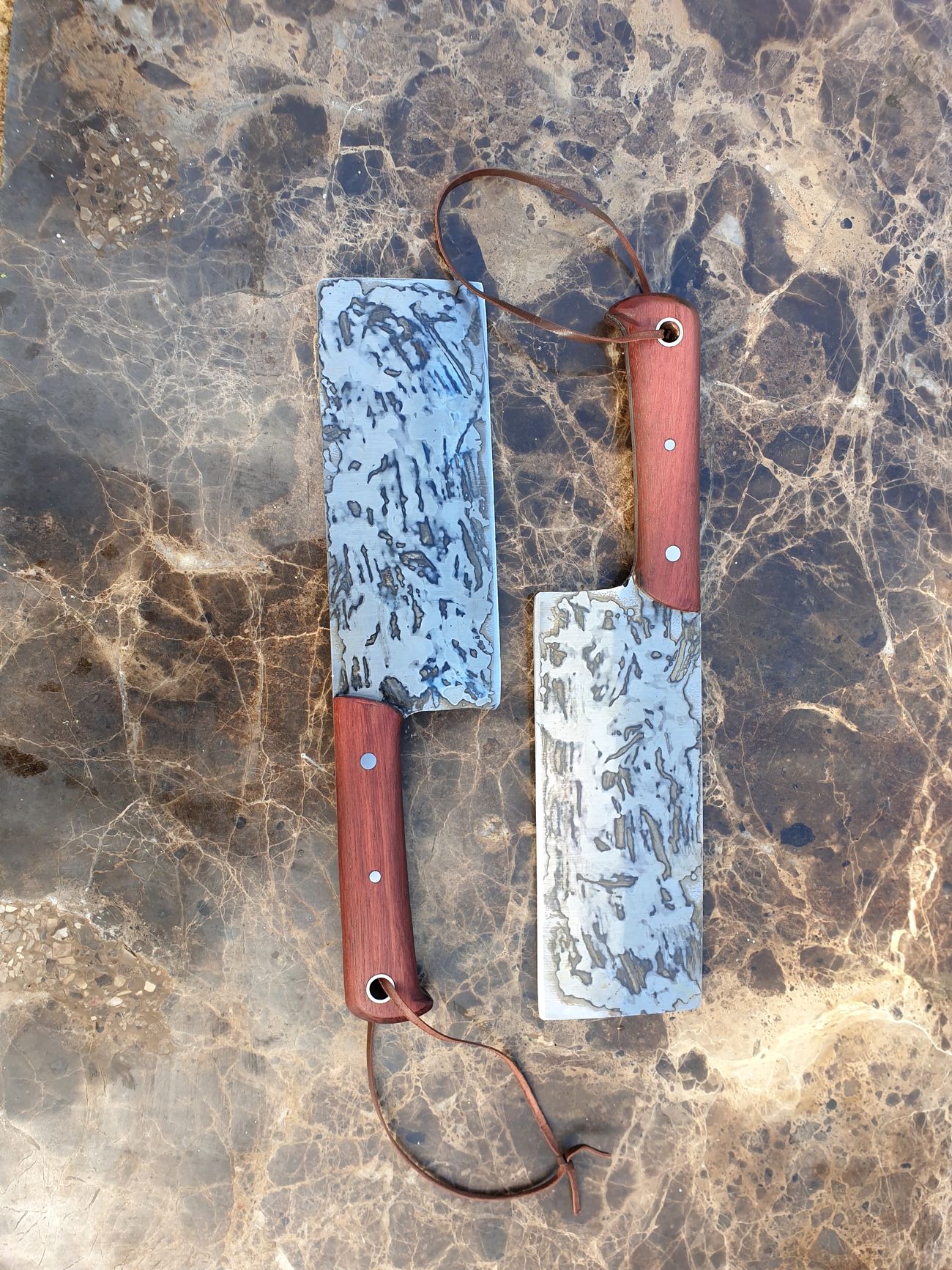 Two handmade cleavers with timber handles and leather lanyards on a marble bench