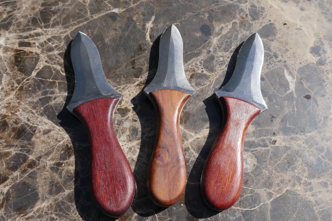 Three finished hand-made oyster knives with different timber handles on a marble bench