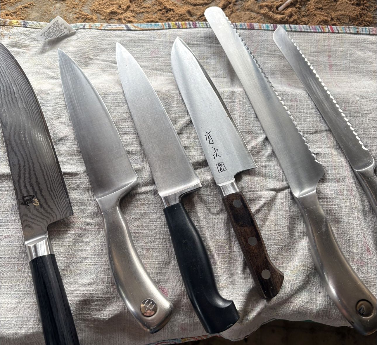 A damascus chef's knife, two chef's knives, a santoku and two bread knives lined up on a cloth after sharpening